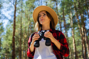 Woman in hat and red plaid shirt holding binoculars in the forest