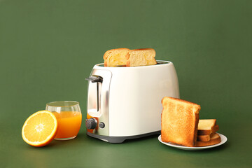 Modern toaster with bread slices and glass of orange juice on green background