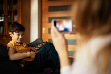 Little boy playing guitar at home. Mother filming her son playing an instrument.