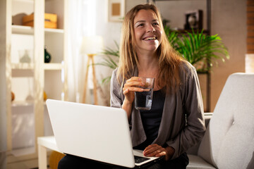 Girl holding a glass of water. Smiling girl drinking water while using the laptop..