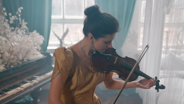 A Young Girl And Man Plays The Violin In A Beautiful Retro Room With Flowers And A Piano. 