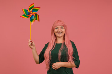 smiling woman with long pink hair in a green dress with a rainbow weather vane in her hands on a pink background