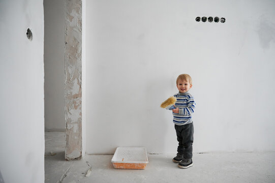 Smiling cute little boy holding special brush for repair in empty room. Concept of process preparing for drawing wall in white color at home during repair.