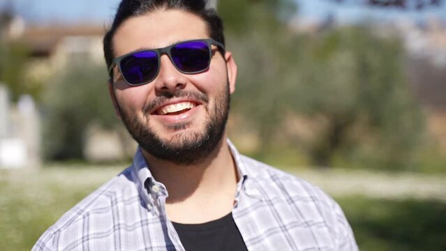 Portrait of an attractive happy bearded casual guy with sunglasses and a checked shirt looking at camera outside.