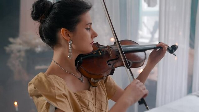 A Young Girl And Man Plays The Violin In A Beautiful Retro Room With Flowers And A Piano.
