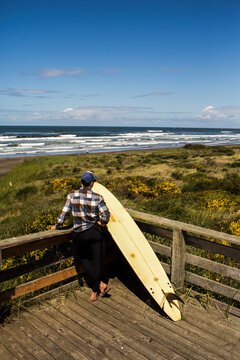 A man with a surfboard overlooks the beach from a deck