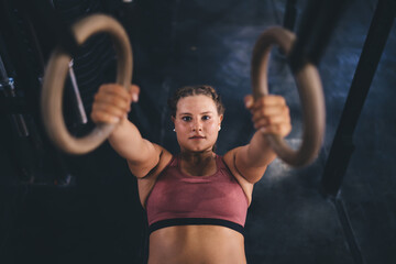 Portrait of Caucasian female athlete performing pull-up on gymnastic rings, motivated fit girl with...