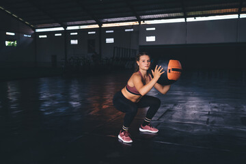 Fototapeta na wymiar Young Caucasian female athlete doing cardio exercises and posing in sportive gym studio, portrait of motivated fit girl with ball equipment looking at camera while squatting during day workout