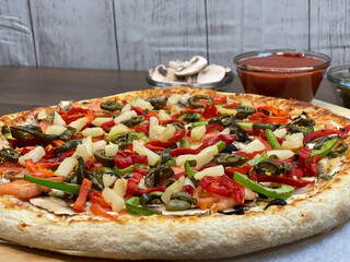 Not cut veggie pizza on wood board on table and pizza cutter and fresh mushrooms and olive oil on side.