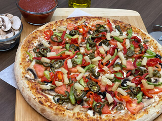 Not cut veggie pizza on wood board on table and pizza cutter and fresh mushrooms and olive oil on side.