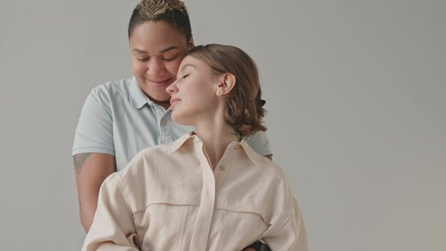 Tilt medium slowmo portrait of tender interracial lesbian couple standing on light background. Masculine Biracial woman embracing her beloved Caucasian girlfriend from behind