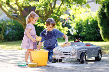 Two happy children washing big old toy car in summer garden, outdoors. Brother boy and little...