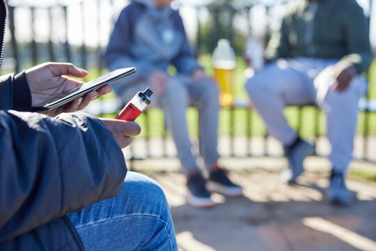 Close Up Of Teenagers With Mobile Phone Vaping and Drinking Alcohol In Park