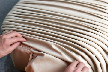 Process of creating folds or draping beige fabric by hand on a furniture pouf. A man pleats fabric...