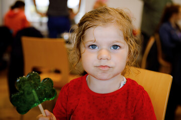 Cute toddler girl with cloverleaf lollipop in Irish pub or cafe. Child resting with parents and...