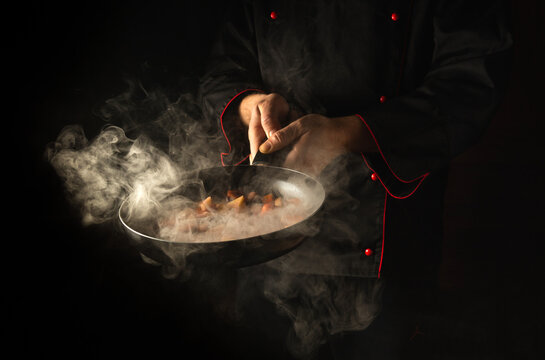 A professional chef prepares food in a frying pan with steam on a black background. The concept of restaurant and hotel service. Vegetarian cuisine.