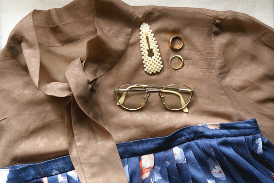 Brown blouse, blue skirt, vintage eyeglasses, gold rings and pearl barrette. Outfit details, flat lay.