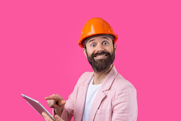 Portrait of young construction engineer wear orange hard hat, in a pink jacket standing on red studio background. A man with a tablet in his hands.
