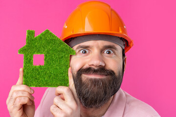 Portrait of young construction engineer wear orange hard hat, in a pink jacket standing on red studio background. A man holds a green eco house.