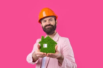 Portrait of young construction engineer wear orange hard hat, in a pink jacket standing on red studio background. A man holds a green eco house.