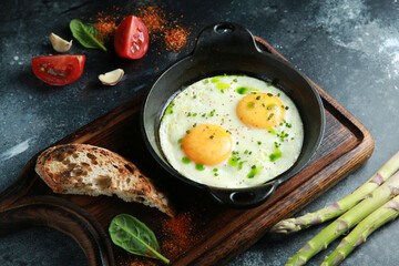 Breakfast. Fried eggs in a cast-iron pan with fried bread, beans, asparagus, tomatoes, herbs on a wooden board on a dark table. Rustic. Background image, copy space.