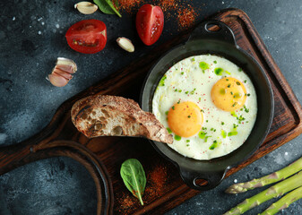 Breakfast. Fried eggs in a cast-iron pan with fried bread, beans, asparagus, tomatoes, herbs on a wooden board on a dark table. Rustic. Background image, copy space. Top view, flatlay