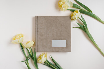 Beige fabric family or wedding photo album with empty space for text surrounded by yellow tulips on a light background. Family photo archive