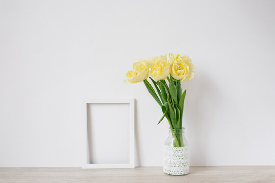 A bouquet of yellow spring tulips in a vase stands on the table, next to an empty white frame for a photo with copy space