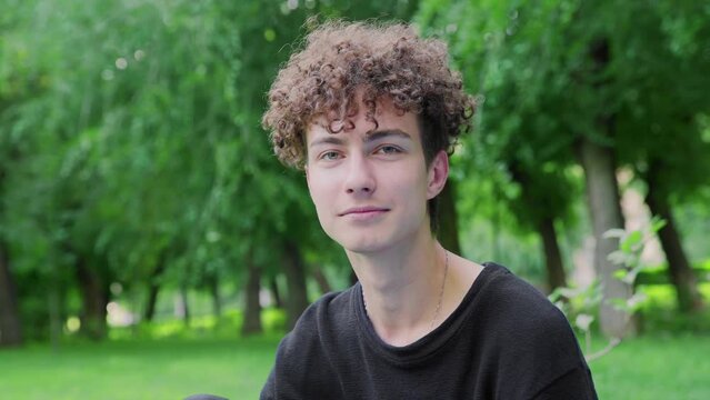 A young curly-haired guy smiles and laughs, looking at the camera in nature in summer
