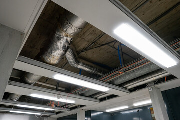 Large pipes mounted on the ceiling of an industrial building corridor. Removed panels to ease access. Neon lights, interior shot, no people.