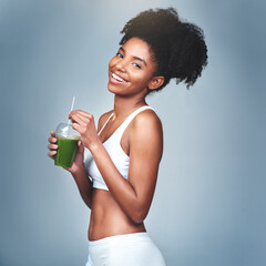I cant go a day without my green juice. Studio shot of an attractive young woman drinking green juice against a grey background. - obrazy, fototapety, plakaty