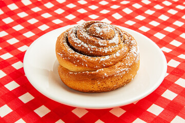 Cinnamon buns or cinnabon rolls, a homemade recipe for sweet traditional Danish dessert with white vanilla powder on red and white checkered background.