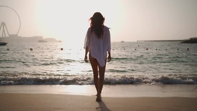 Woman in a white shirt walks along the beach of the Persian Gulf in Dubai towards the sea against the backdrop of a beautiful sunset. Journey through the United Arab Emirates. Slow motion. 
