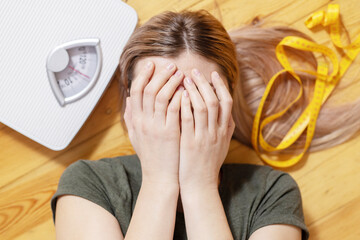Upset and sad woman covering face with hands while lying on wooden floor with white scales and...