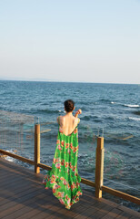 Asian woman in maxi green floral print dress, looking over the ocean from a bar balcony