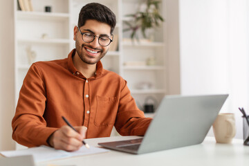 Smiling Arab man in glasses using laptop and taking notes