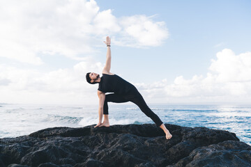 Calm male in sportive wear standing in yoga pose and meditate during morning mindfulness concentration at coastline seashore, Caucasian man enjoying hatha practice for enlightenment harmony