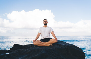 Portrait of calm male in sportive wear sitting in lotus pose and looking at camera during morning meditation near ocean, young Caucasian man relaxing during yoga concentration at nature coastline