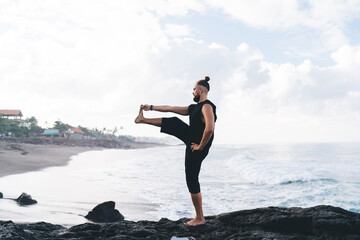 Side view of pumped up male yogi recreating doing balance yoga poses feeling inner peace and vitality, male with strong body standing in asana care about physical and mental health at seashore