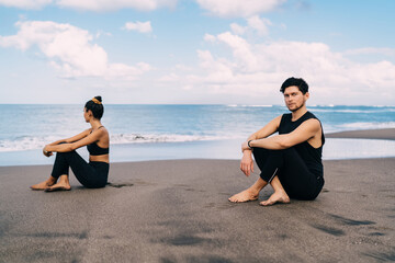 Portrait of Caucaisan male in sportive clothes resting and meditate at seashore beach during time for workout training, young man looking at camera during together retreat healing with female friend