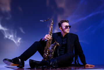 Side view of handsome male musician, wearing in trendy black suit and sunglasses, sitting on floor...