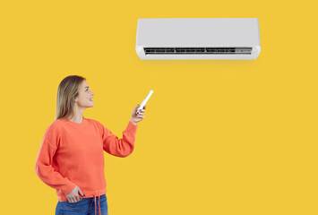 Happy girl turns on the AC. Young lady switches on the AC on the wall. Beautiful woman standing isolated on a yellow colour background setting comfortable temperature her air conditioner at home