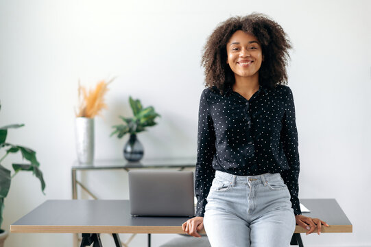 Successful happy pretty young dark-skinned girl, business woman, broker, financial consultant, stands near the desktop in the office, dressed in stylish clothes, looks at the camera, smiles friendly.