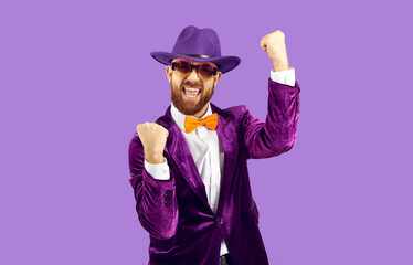 Happy confident man celebrating success. Funny excited cheerful bearded guy in hat, suit and orange bow tie standing on purple background, fist pumping and shouting Yes, Yay, I did it, Cool, Hurray