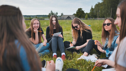 A group of female students are sitting in a circle on a meadow for collective work with notebooks.