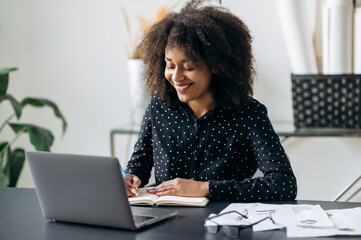 African american company manager or freelancer working in the office. Attractive confident smart curly haired woman, sitting in front of a laptop, making notes in a notebook, listens webinar, smiling