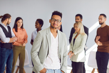 Fototapeta na wymiar Young black man in suit jacket, casual tee and stylish glasses standing hands in pockets in front of diverse coworkers. Portrait of handsome businessman, team leader or business conference participant