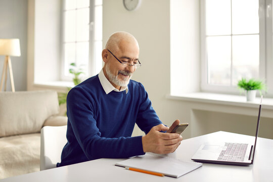 Man sitting at table with mobile phone. Happy bald bearded senior man in glasses sitting at desk with laptop and notepad, using modern cellphone, reading news, looking up and studying new information