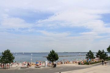 View of beach in Kiel, Germany with crowd of people sunbathing and swimming in summer with clouds in blue sky background.