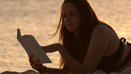 Young woman reads a book while lying on the beach at sunset